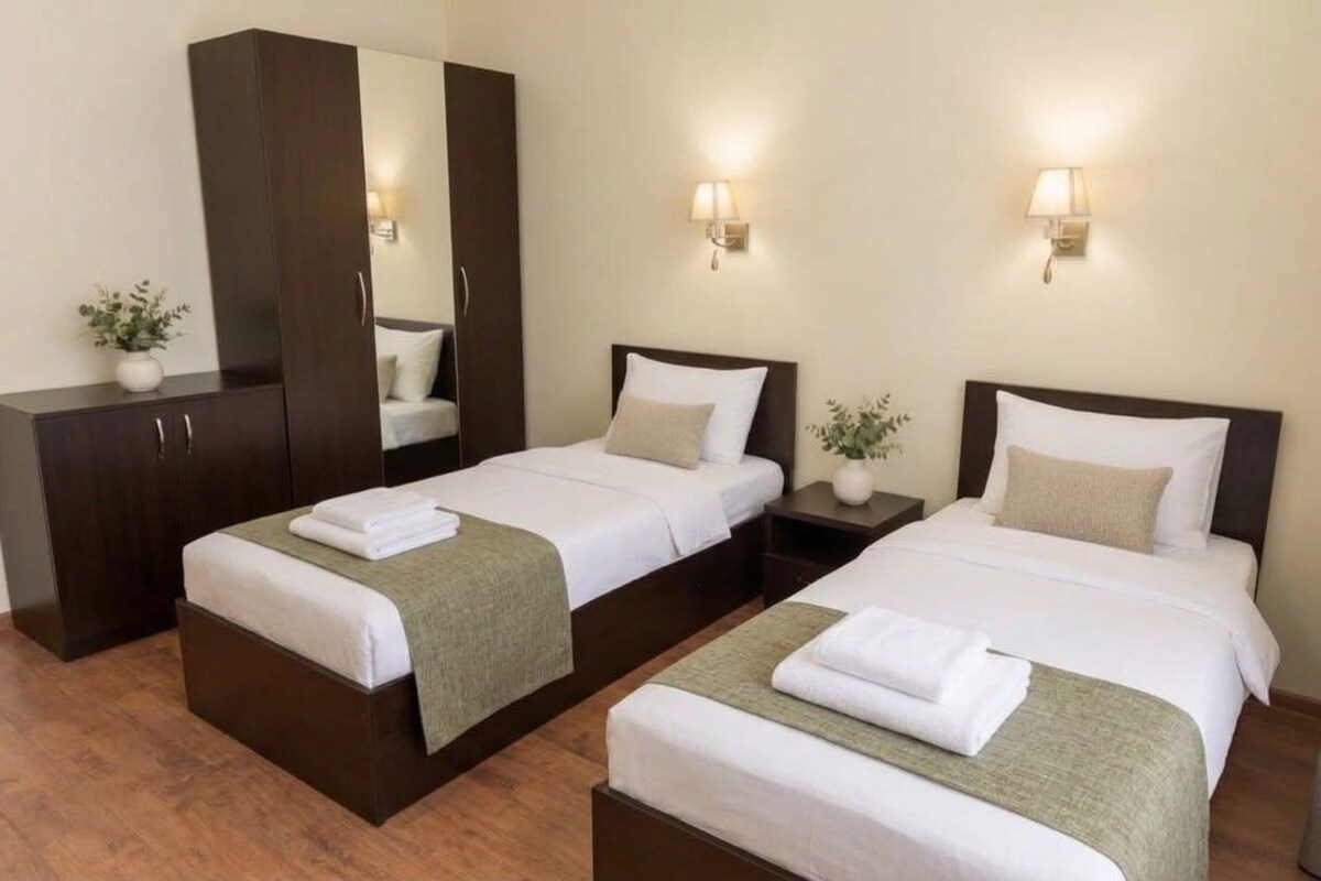 A hotel room with two single beds, a wardrobe, bedside lamps, and a small potted plant.