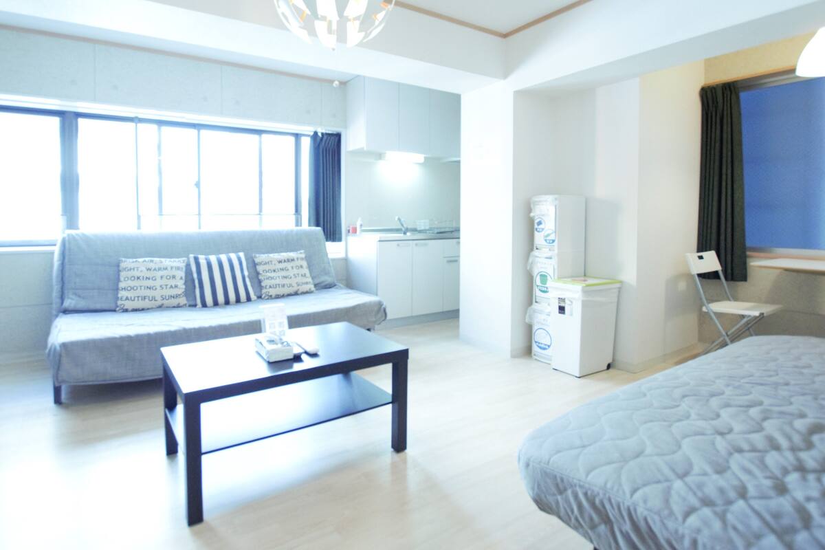 Una habitación de hotel moderna con sofá, cama, una mesita, kitchenette y ventana con cortinas.
