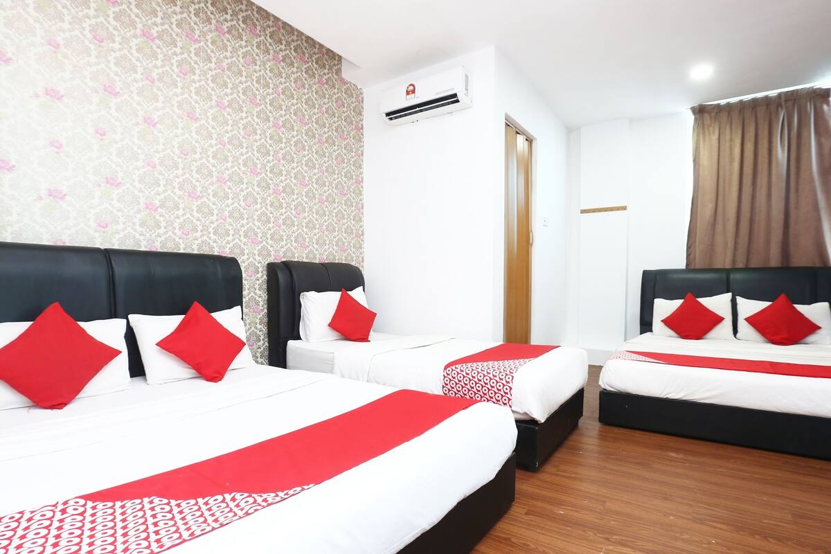 A hotel room with two beds, each with a black headboard and red and white bedding. There is a floral-patterned wallpaper on the wall, a wooden floor, and a window with brown curtains.