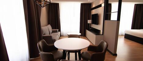 A modern hotel room with a round dining table, four chairs, a sofa, a TV mounted on the wall, and a large window with curtains.