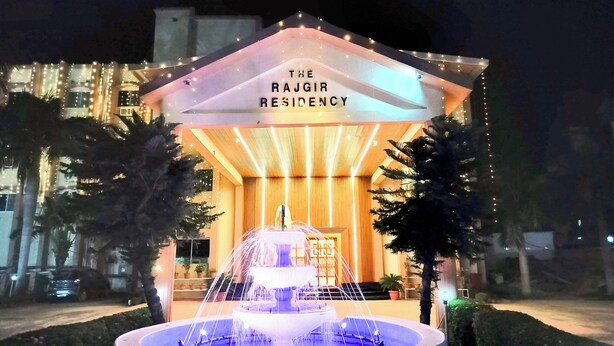 El Rajgir Residency con una fuente en la entrada, rodeado de plantas en macetas y palmeras.