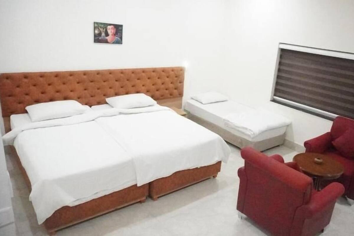 Une chambre d’hôtel avec un grand lit, un lit plus petit, un fauteuil rouge, une petite table et une fenêtre avec des stores.