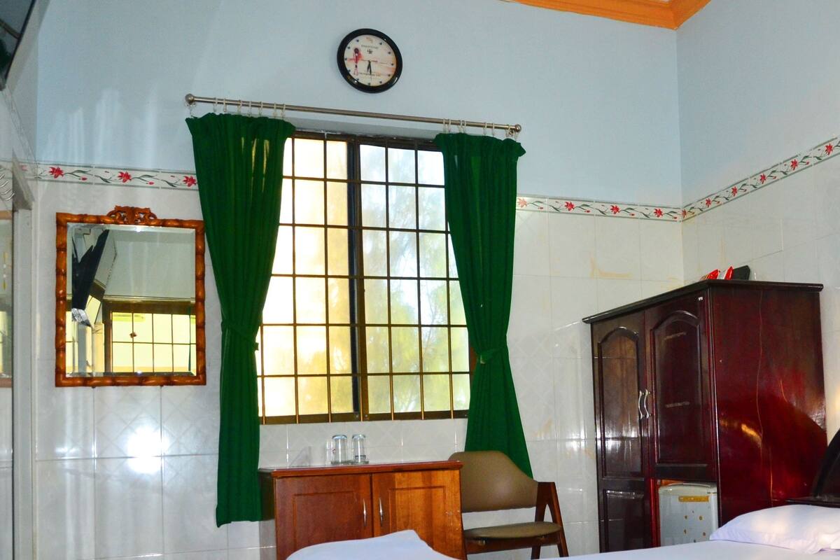Une chambre d’hôtel avec un lit, une armoire en bois, une chaise, une petite table, un miroir et une fenêtre avec des rideaux verts.