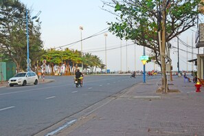 Une rue où une voiture blanche est garée sur le bord, une moto avec son conducteur, et un trottoir pour les piétons.