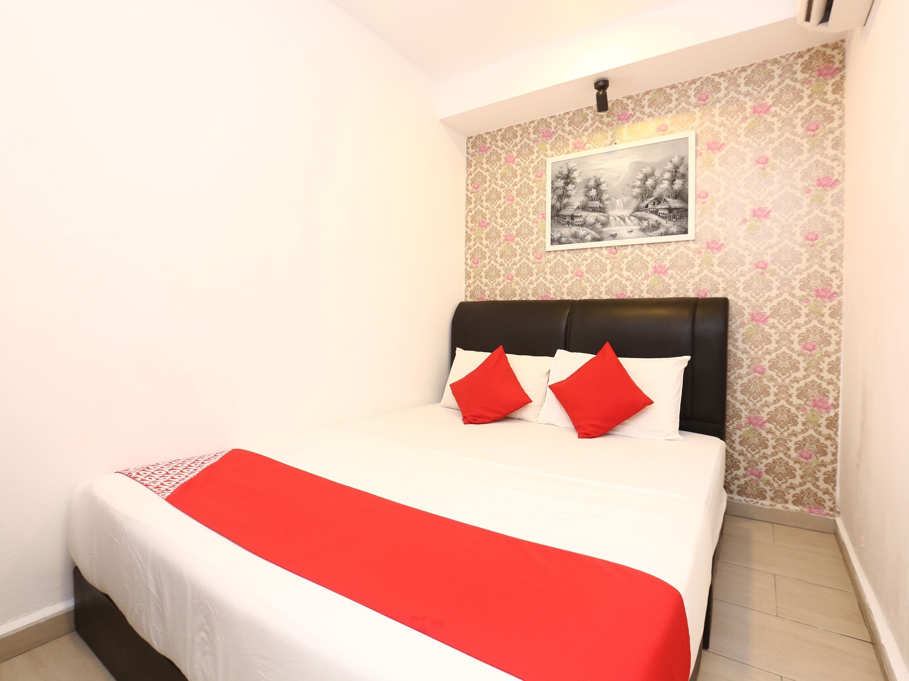 A neatly made bed with a red bedspread and pillows, a black headboard, and a patterned wallpaper with a black and white framed picture above it.