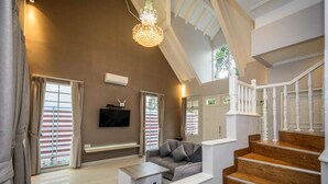 A spacious living room with a grey sofa, a coffee table, and a TV mounted on the wall. There is a staircase with wooden steps and white railings leading to an upper level. - Villa Kota Bunga Puncak 2 Bedroom (Cipanas)