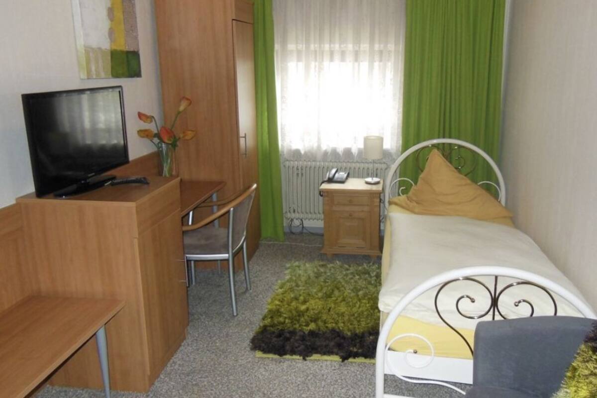 A bedroom with a bed, a desk with a chair, a television, a radiator, and a window with curtains.