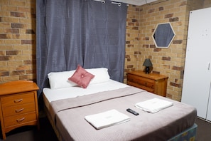 A hotel room with a brick-patterned wall, a bed with a white and pink pillow, a wooden nightstand, a dresser, and a white wardrobe. - Fantasy Ocean Holiday House (Coffs Harbour)