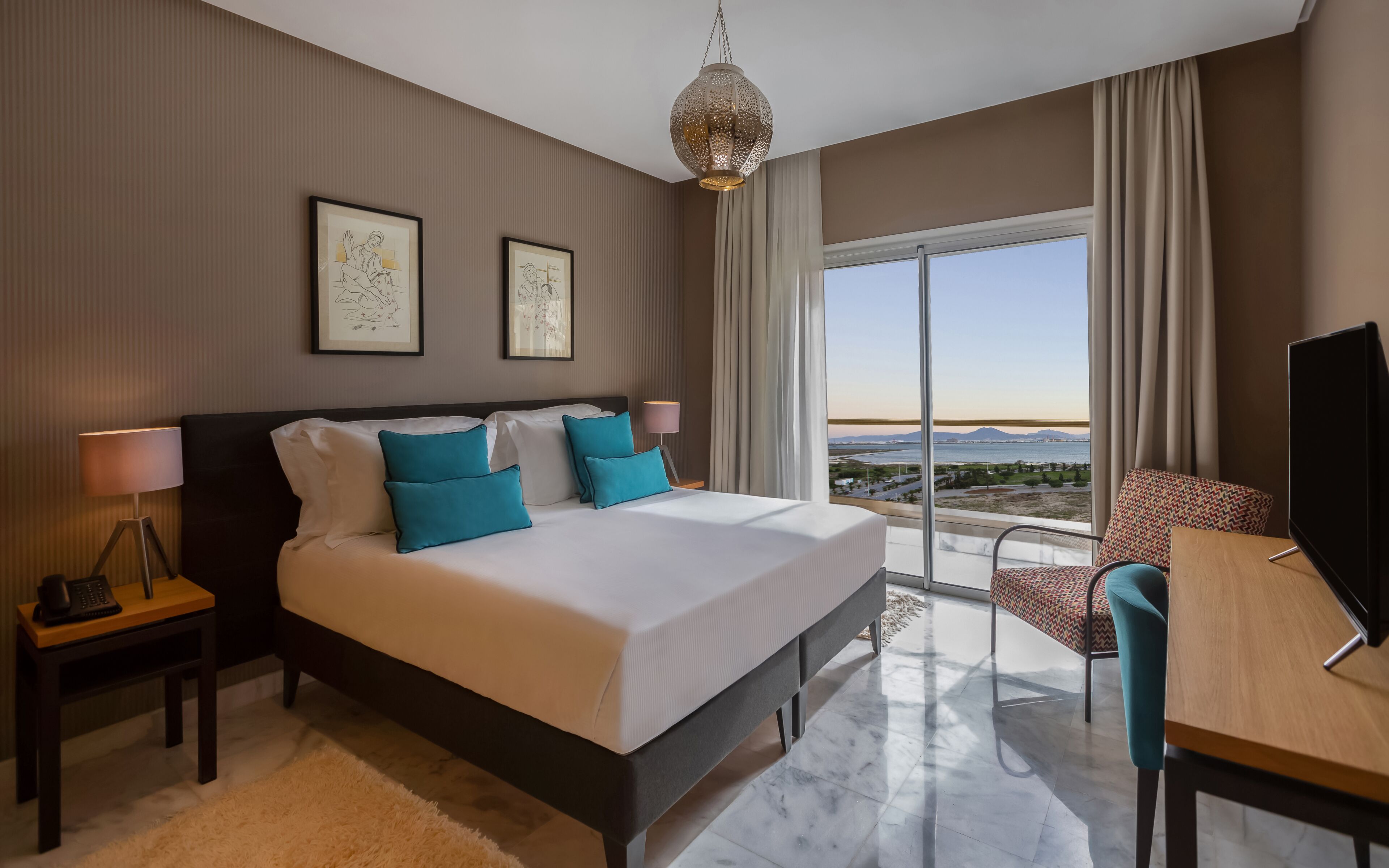Quarto de hotel com uma cama grande, uma escrivaninha, uma cadeira, uma televisão, um abajur e vista para o mar.