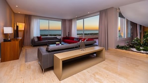 Habitación de hotel moderna con un sofá grande, una mesa de centro y un escritorio con lámpara. Amplias ventanas ofrecen vistas al mar.
