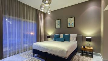 A modern bedroom with a large bed, two bedside tables with lamps, a large window with sheer curtains, and two framed artworks on the wall.