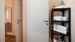 Uma casa de banho moderna com uma porta branca, uma prateleira de madeira com toalhas e uma pequena planta no chão.