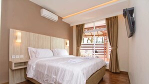 A bedroom with a wooden headboard, a bed with white linens, an air conditioning unit, a TV mounted on the wall, and a window with curtains. - Villa Kota Bunga Puncak 4 Bedroom (Cipanas)
