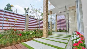 A modern outdoor area with a striped wall, a glass door entrance, and a pathway with artificial grass and flowers. - Villa Kota Bunga Puncak 4 Bedroom (Cipanas)