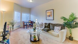 A living room with a sofa, coffee table, and a dining table with chairs. There is a large window with blinds, a framed picture on the wall, and a potted plant. - Cosy Berkeley Apartments Across UC Berkeley #121 (Berkeley)