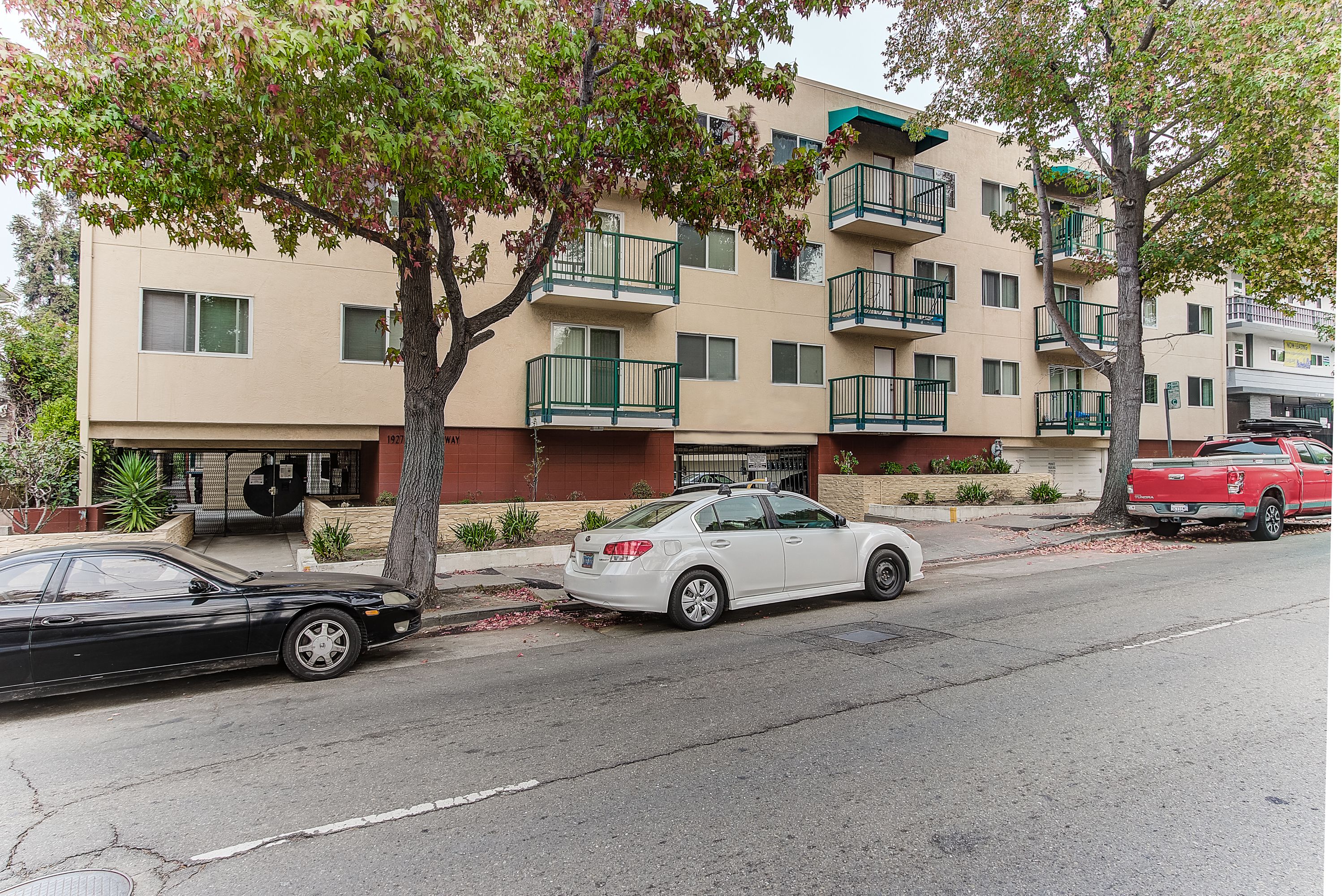 A multi-story residential building with balconies, a parked white car, and a red truck on the street.