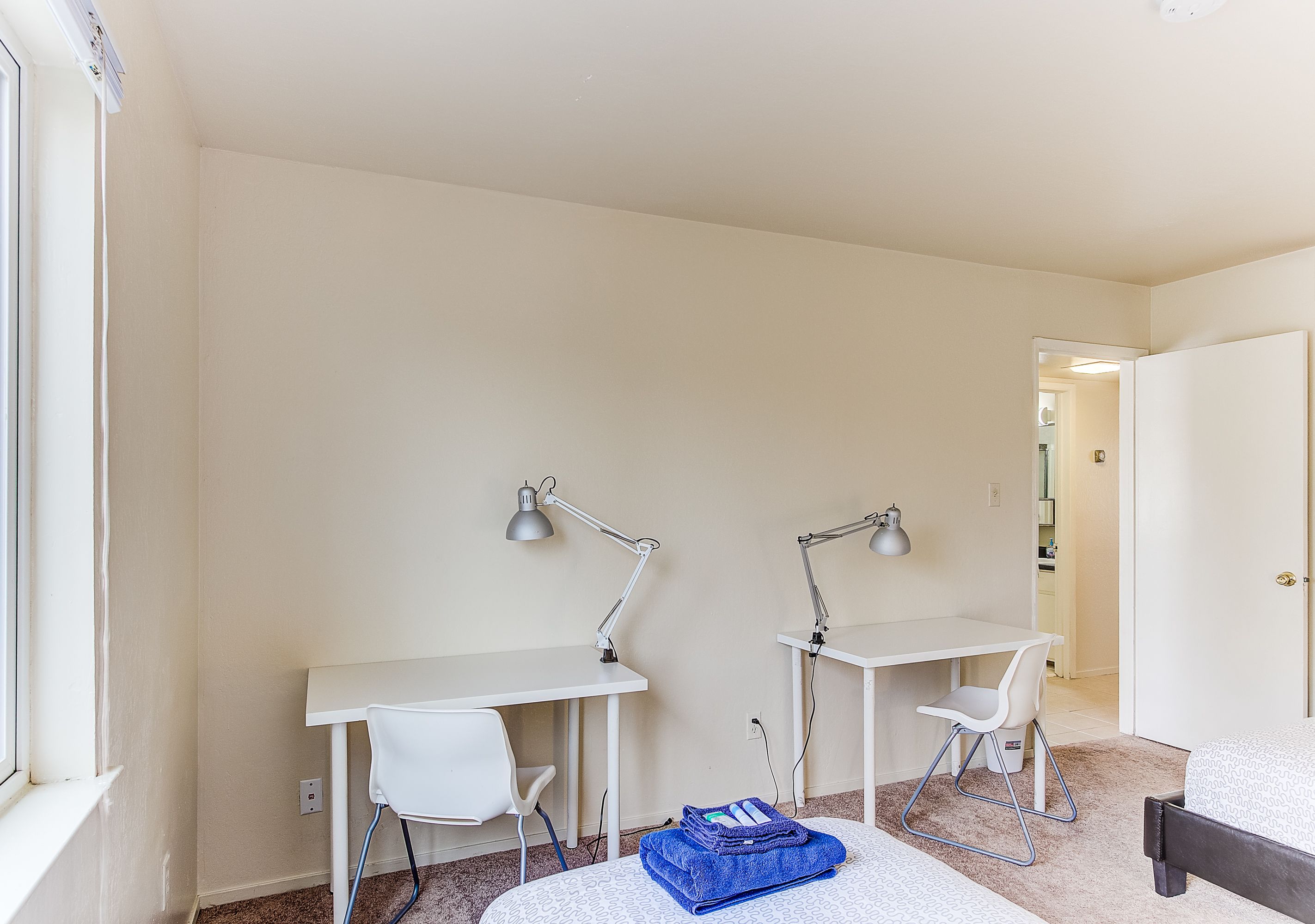 Una habitación con dos escritorios blancos, cada uno con una lámpara, una silla blanca y un bolso azul en el suelo.