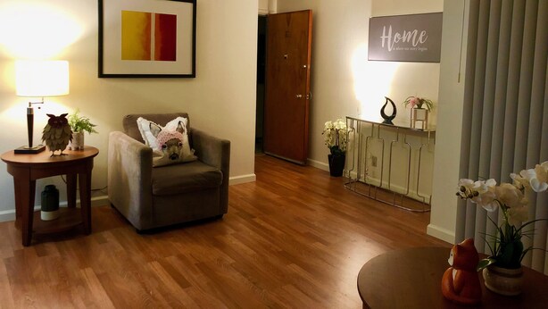 A living room with a wooden floor, a grey armchair with a pillow, a side table with a lamp, a framed abstract painting, a 'Home' sign, a door, and a small table with a flower vase. - Cosy Berkeley Apartment Close to UC Berkeley #E (Berkeley)