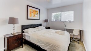 Quarto com cama, duas mesas de cabeceira com luminárias, escrivaninha com cadeira, janela com persianas e ventilador de teto.