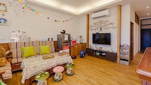 A cozy living room with a sofa, a coffee table, and a TV mounted on the wall. There are colorful decorations on the wall and a small table with a basket.