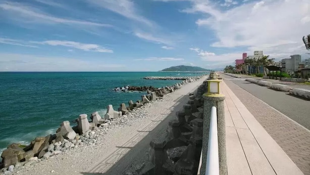 A coastal walkway with a rocky shoreline, a clear sky, and buildings in the background.