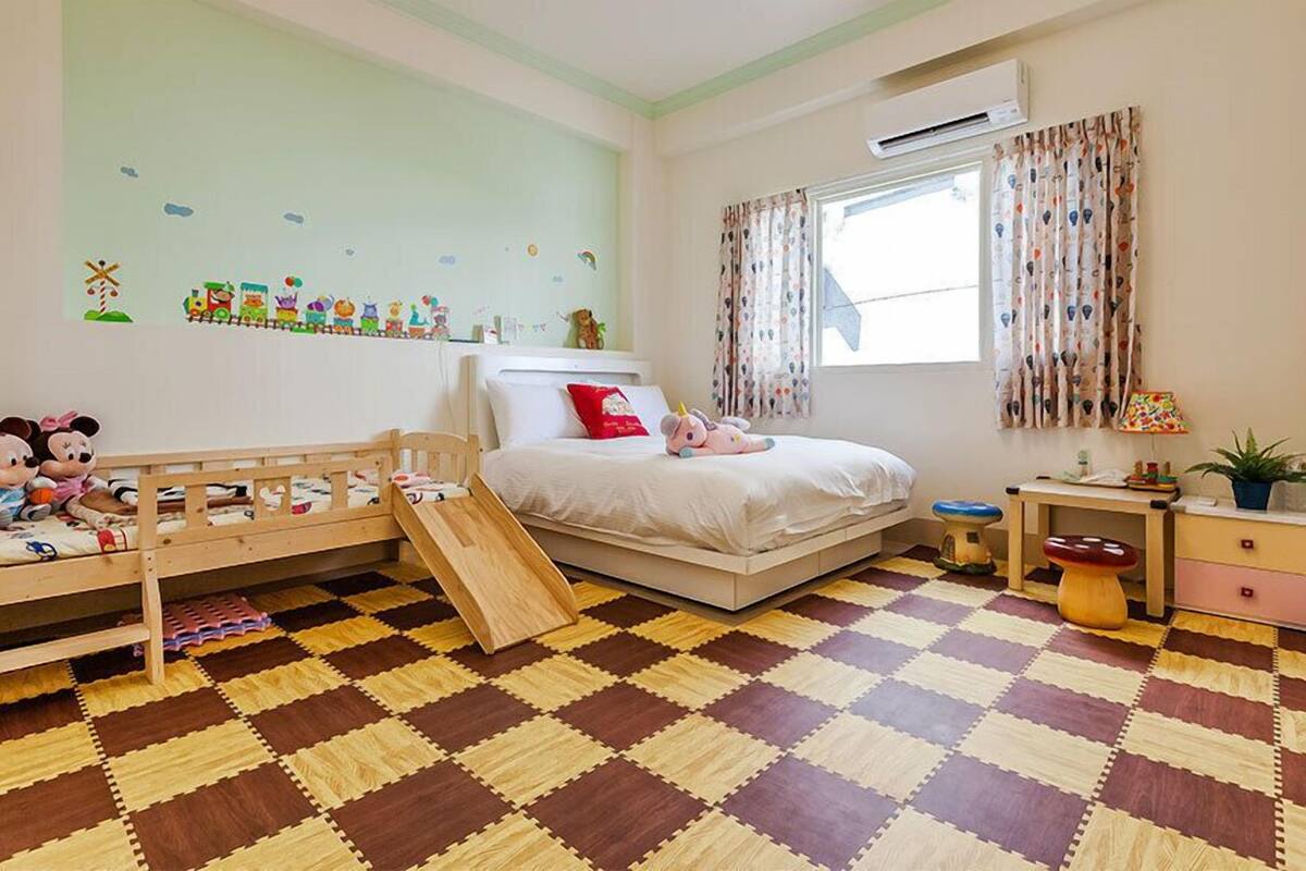 A child's bedroom with a bed, a wooden playpen, a small table, and a window with curtains.