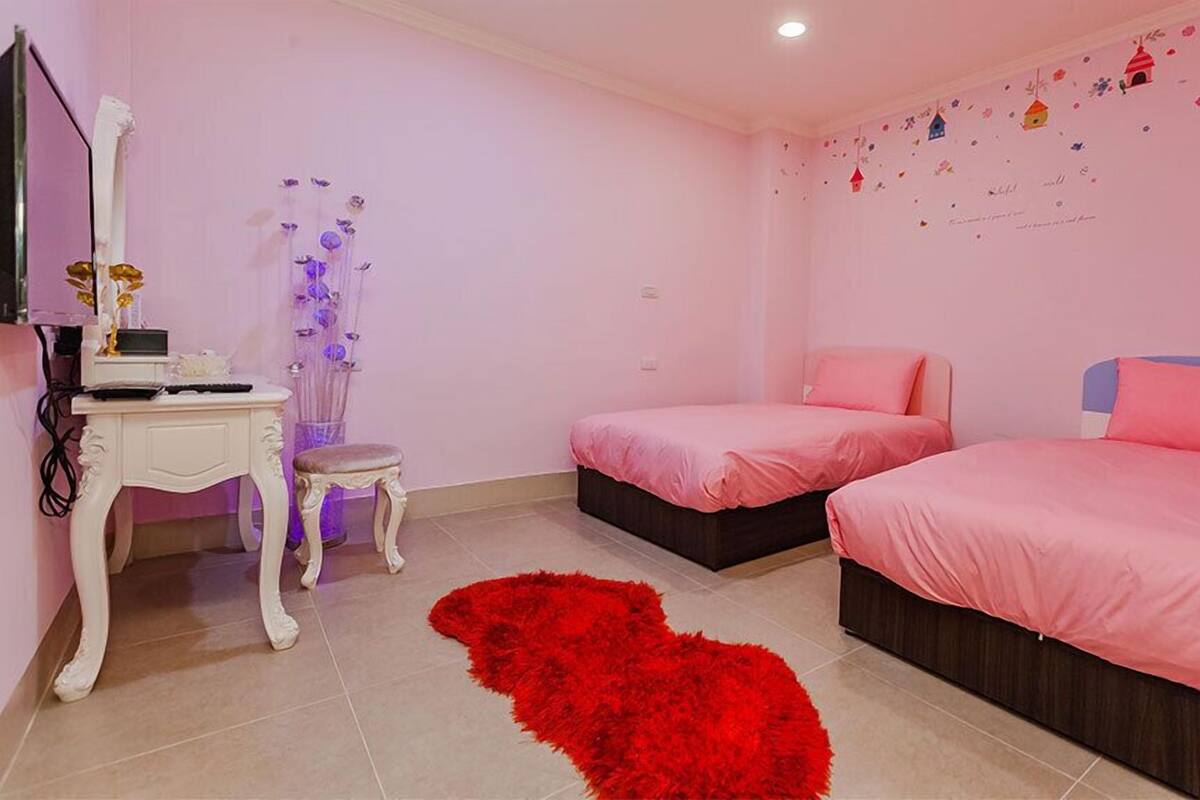 A pink-themed bedroom with two beds, a desk, and a red rug.