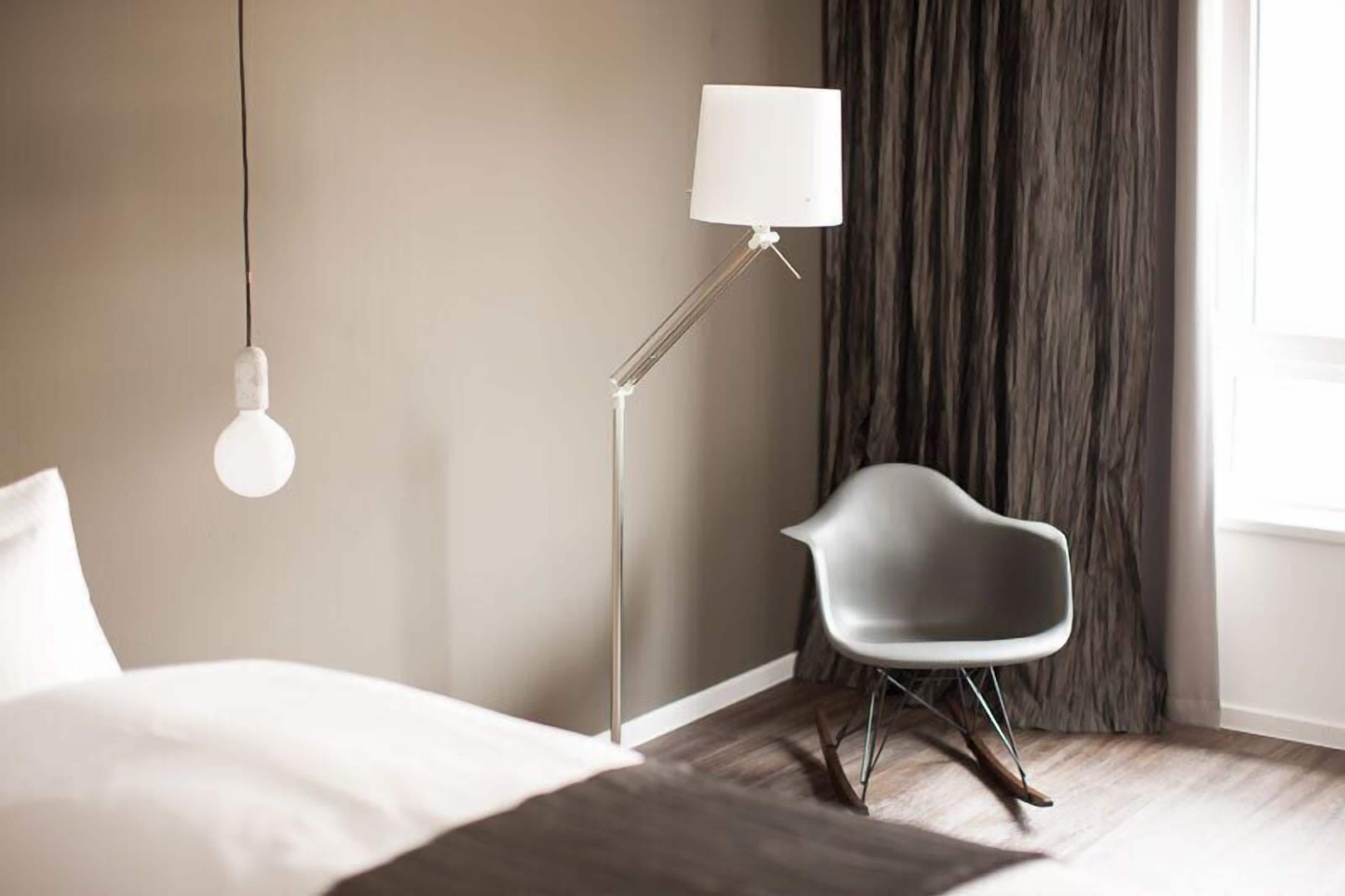 Un dormitorio moderno con una cama, un sillón gris, una lámpara de pie y una ventana con cortinas.
