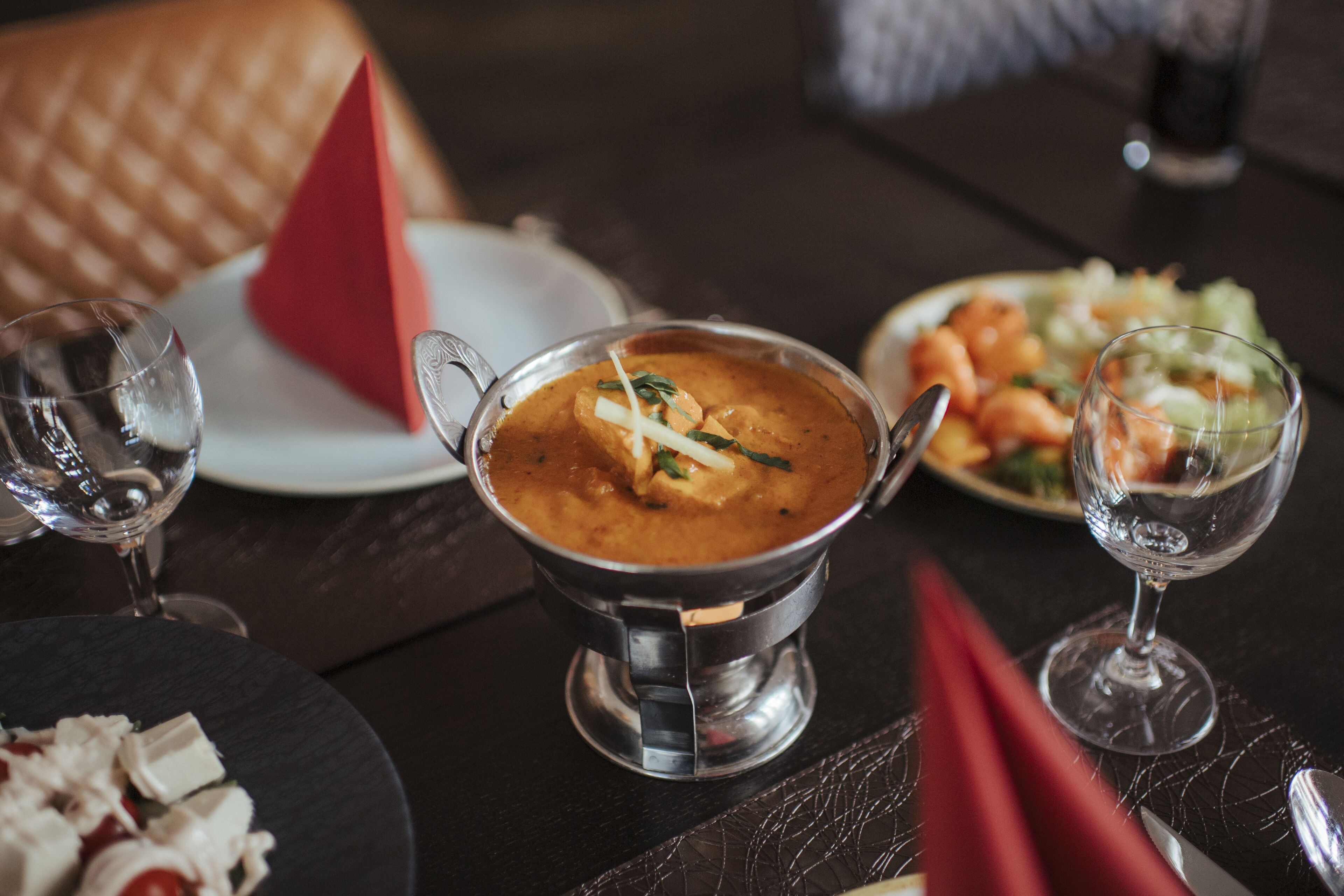 A dining table with a metal bowl of soup, a plate of shrimp, and a red napkin.