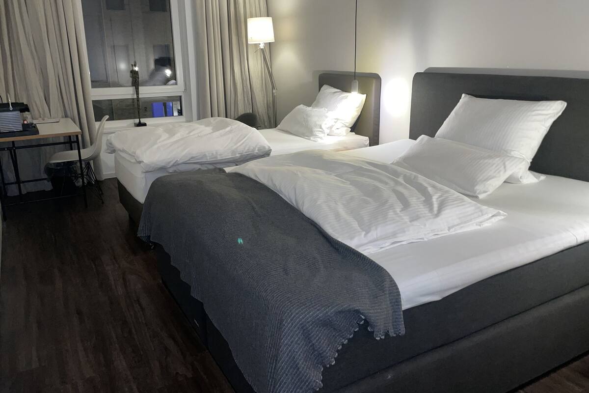 Ein Hotelzimmer mit zwei Betten, einem Schreibtisch, einem Stuhl, einer Lampe und einem Vorhangfenster.