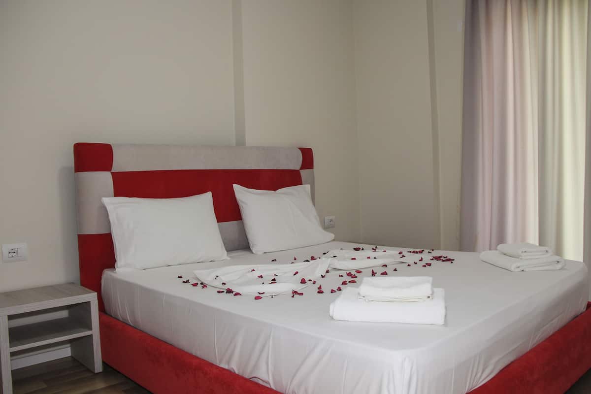 Un lit bien fait, avec du linge de lit blanc et des touches de rouge, une table de chevet et une fenêtre avec des rideaux.