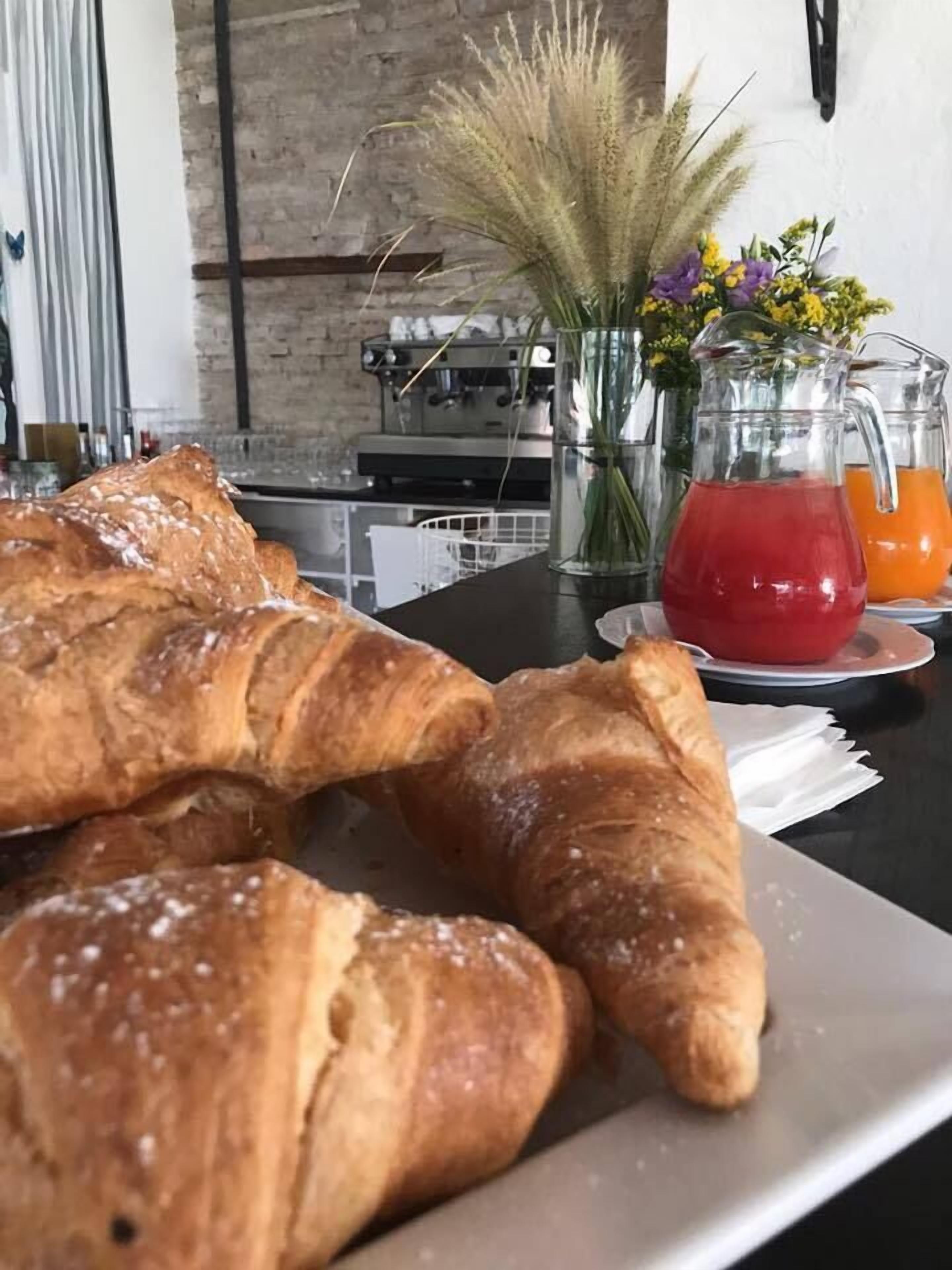 Taula amb croissants i una gerra de beguda vermella, amb una paret de maó i electrodomèstics de cuina al fons.
