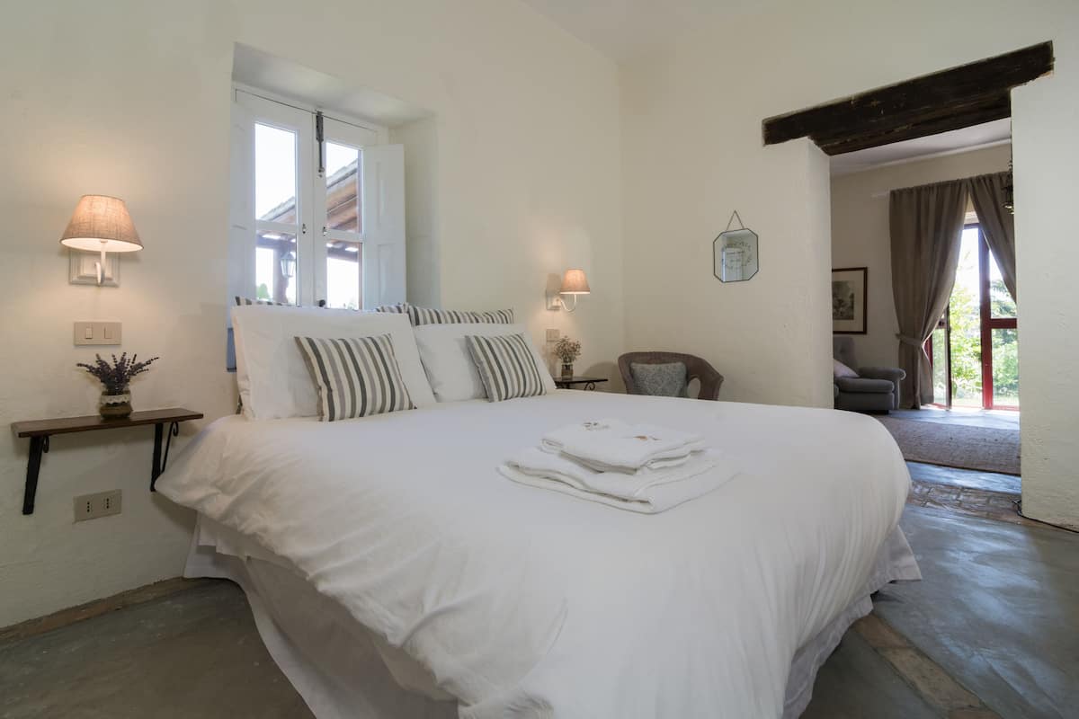 A large bed with white linens, two bedside lamps, a small table with a plant, and a window with curtains.