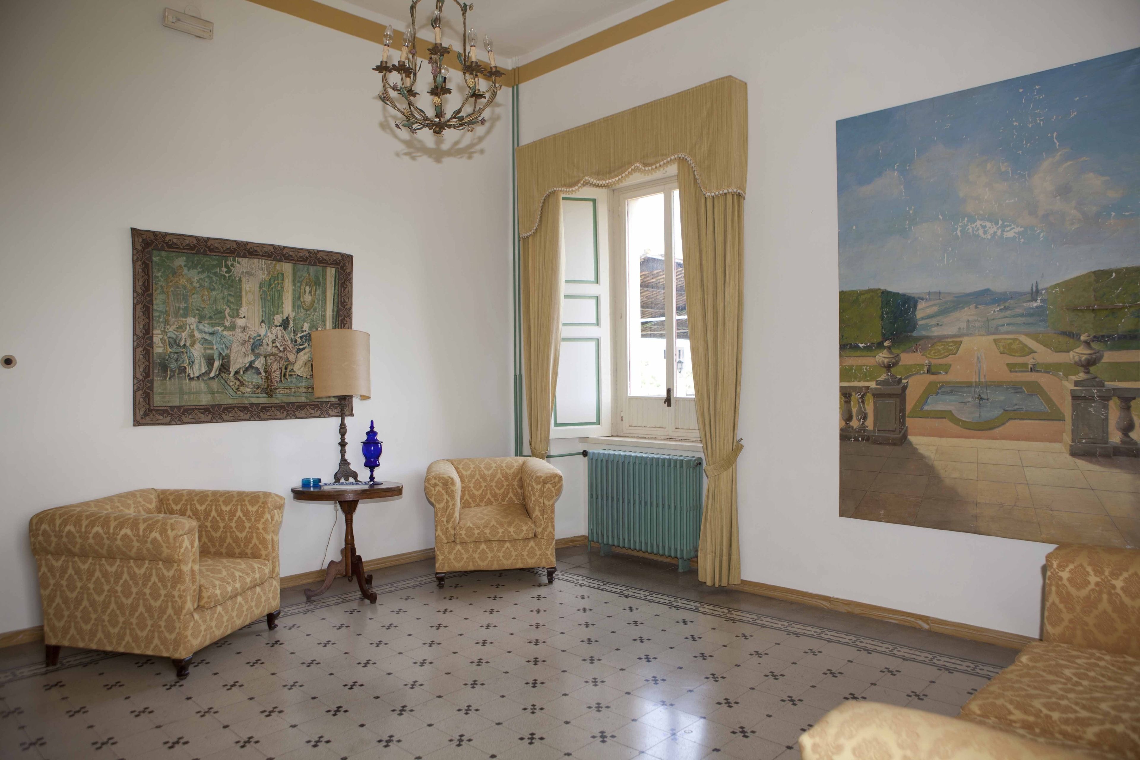 A room with patterned floor tiles, two armchairs, a table with a lamp, a painting on the wall, a window with curtains, and a large landscape painting.