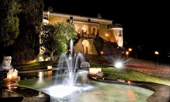Vista nocturna de un edificio histórico con una fuente, iluminado por luces y rodeado de árboles.