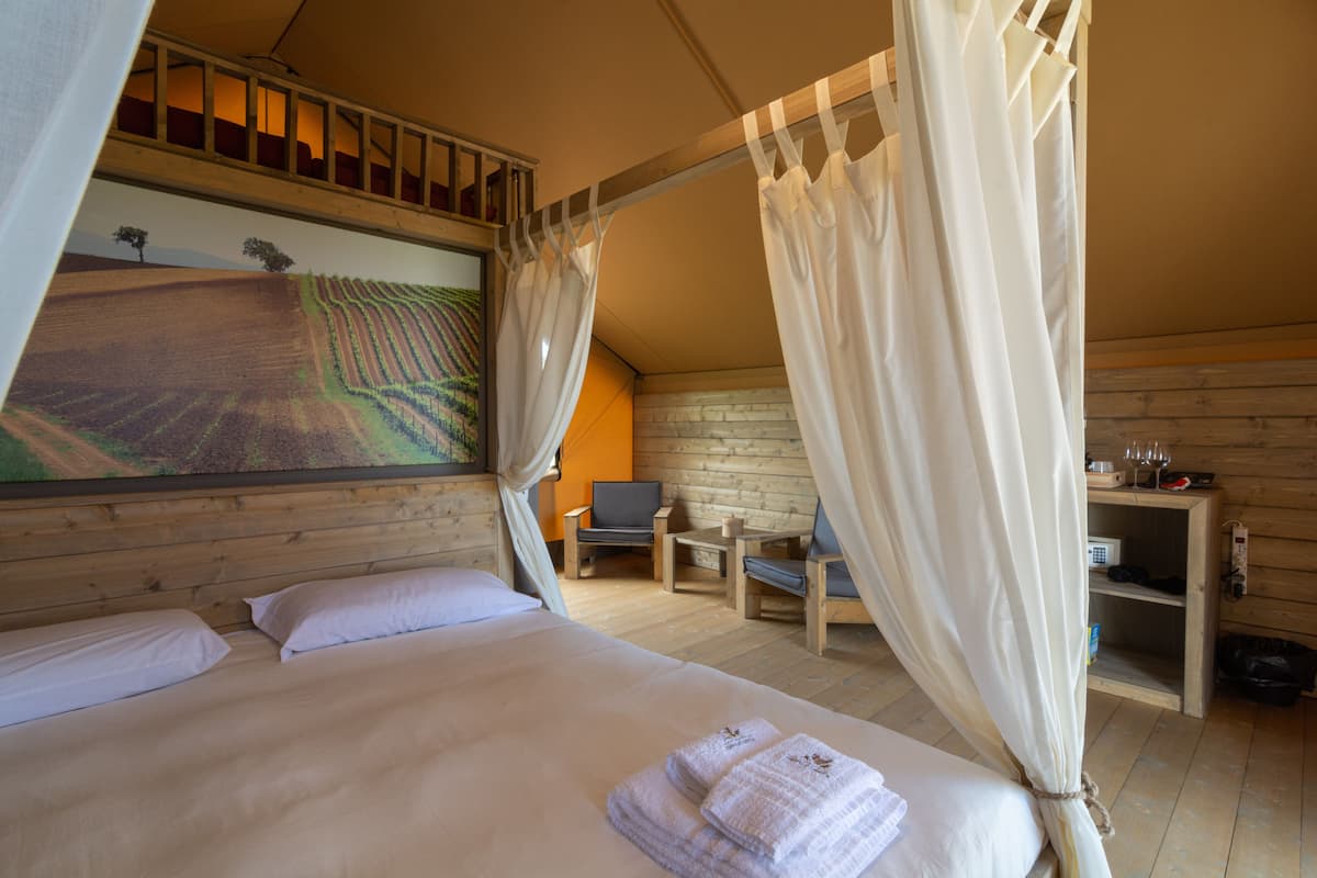 A bedroom with a large bed, a desk, and a chair. There is a painting on the wall and a window with curtains.