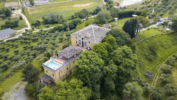 Une vue aérienne d’un grand bâtiment jaune doté d’une piscine, entouré d’une végétation luxuriante et d’un paysage vallonné.