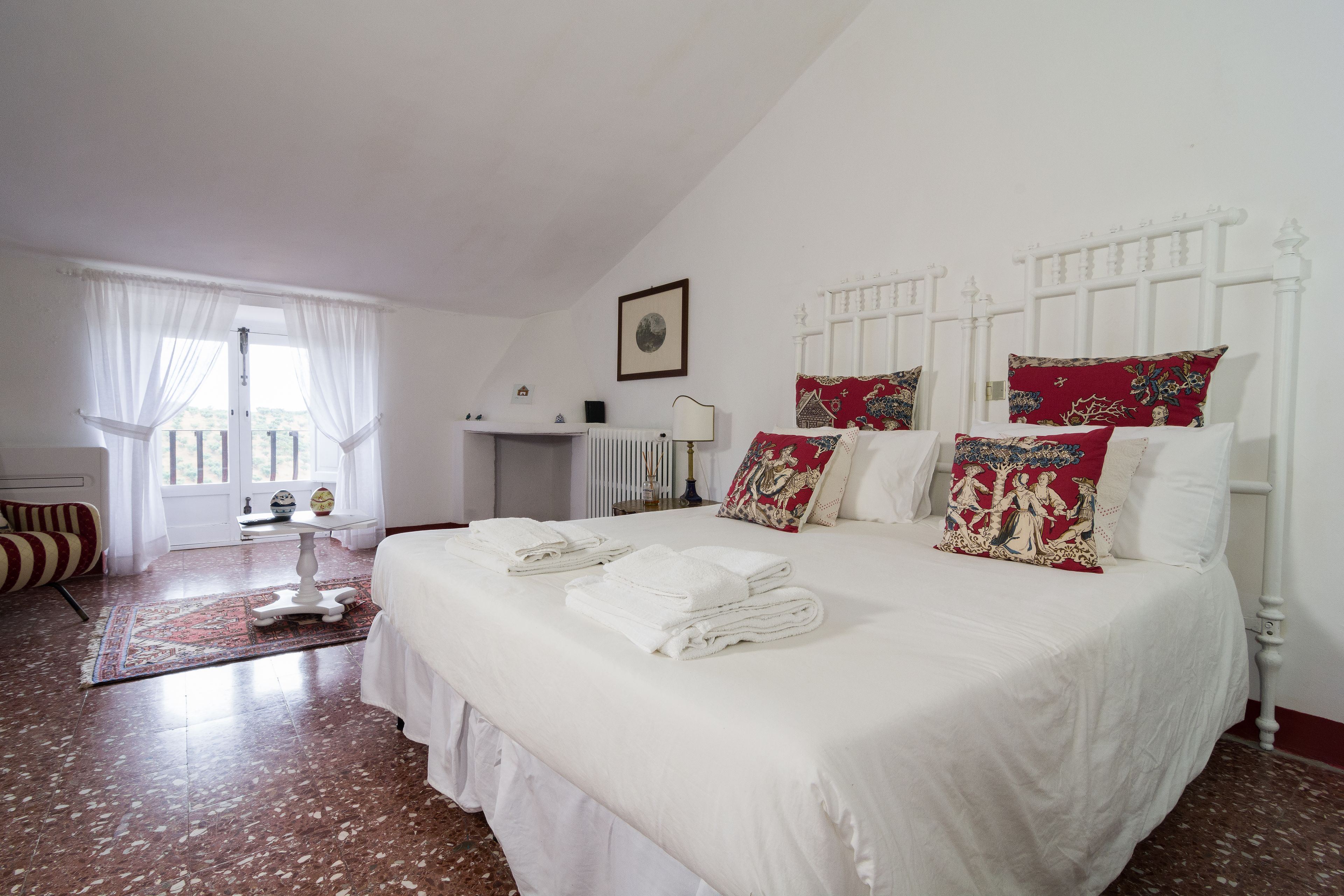 Una cama bien tendida con ropa blanca y toallas, un radiador blanco, una mesita con una lámpara y una ventana con cortinas translúcidas.