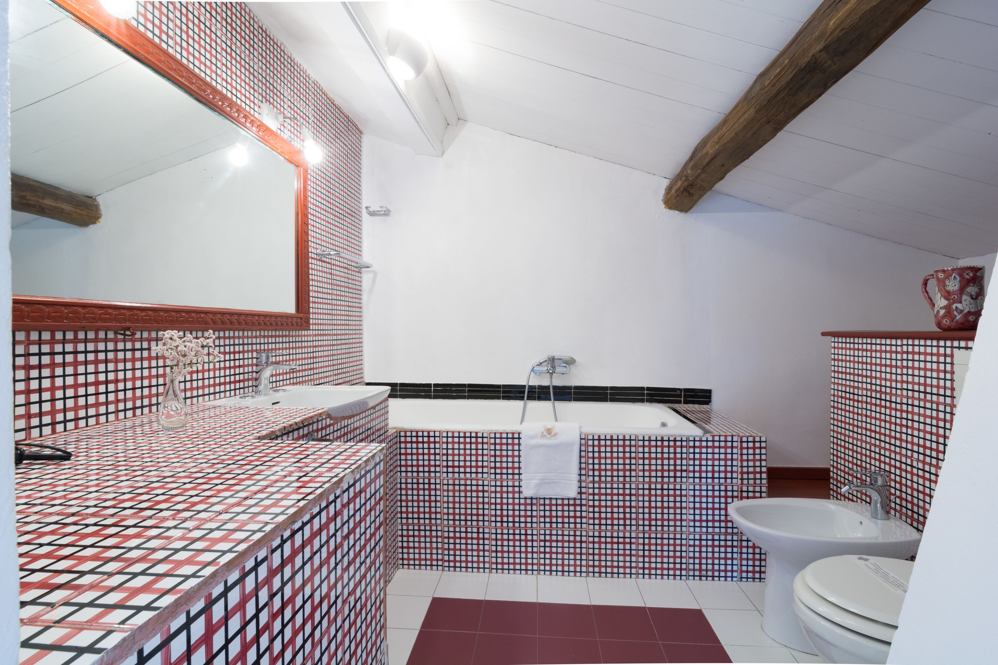 A bathroom with a red and white checkered tile pattern, a large mirror, a bathtub, and a toilet.