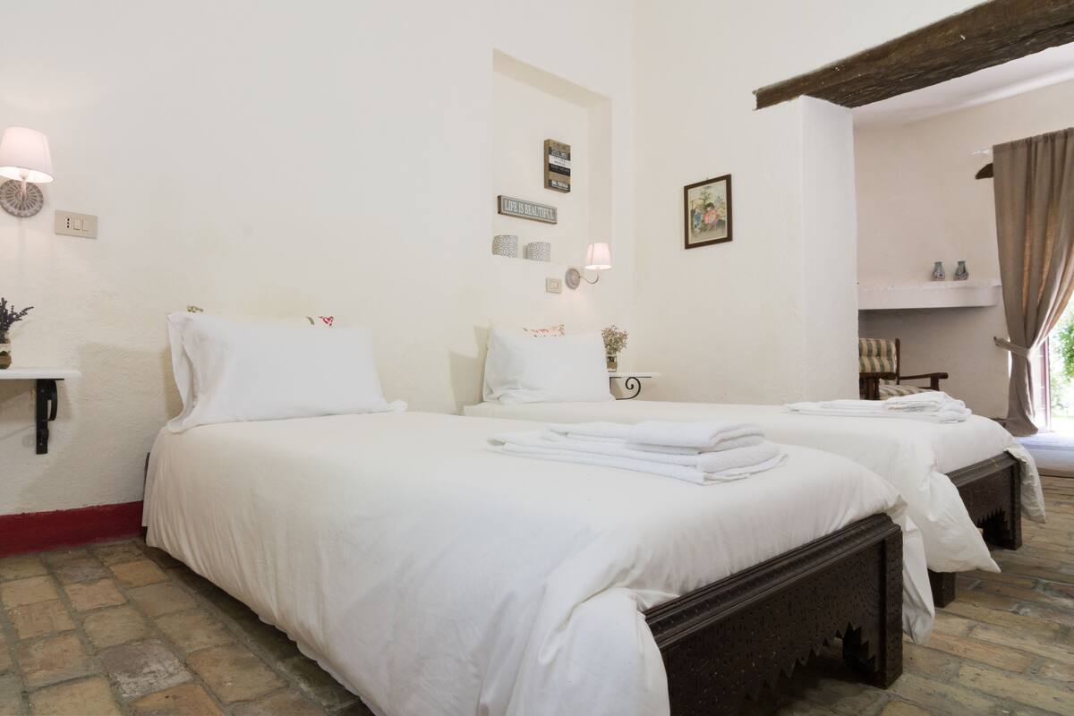 A bedroom with two single beds, a bedside table with a lamp, a wall clock, and a small shelf with decorative items.