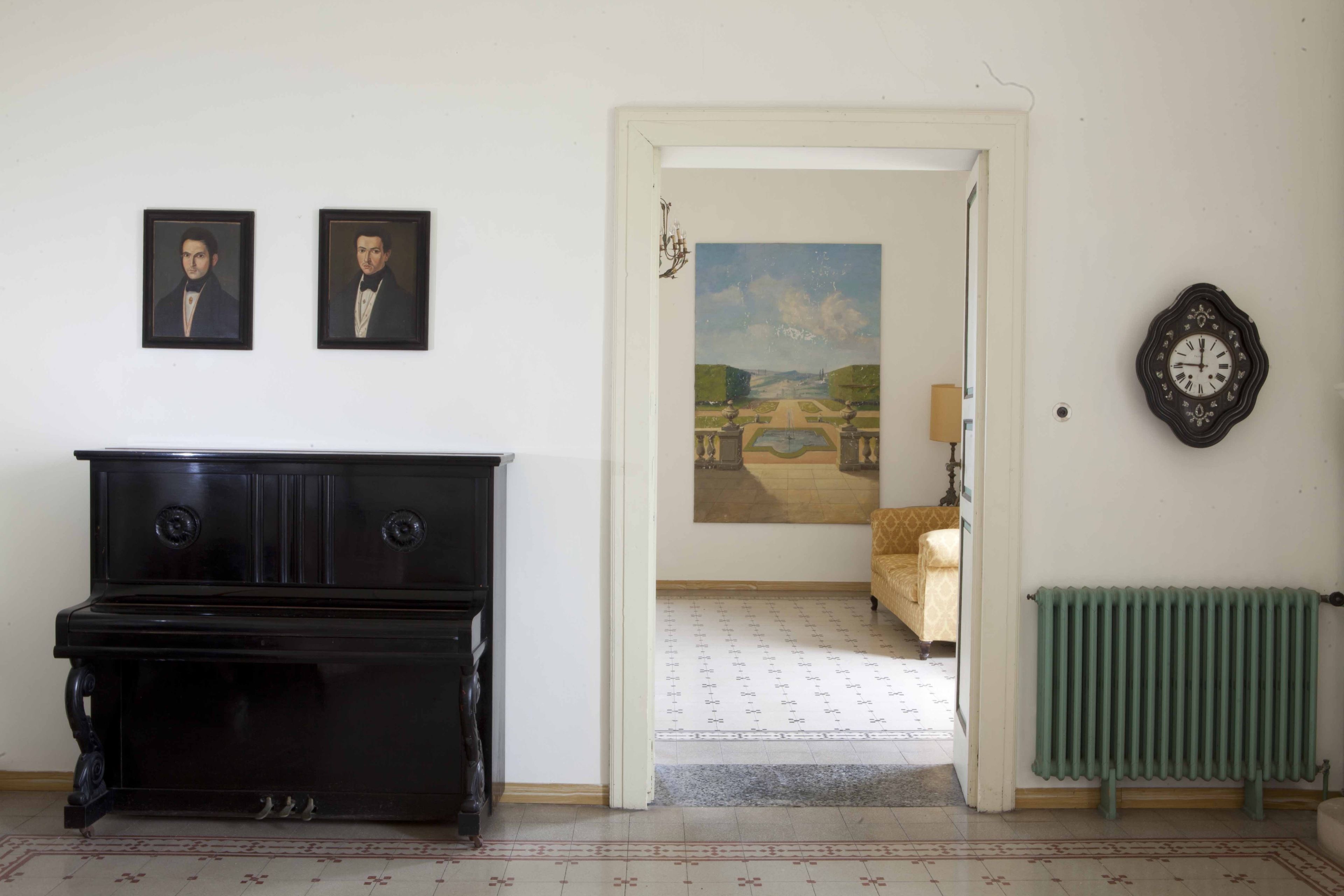 A room with a black piano, two framed portraits on the wall, a painting of a landscape, a radiator, and a clock.