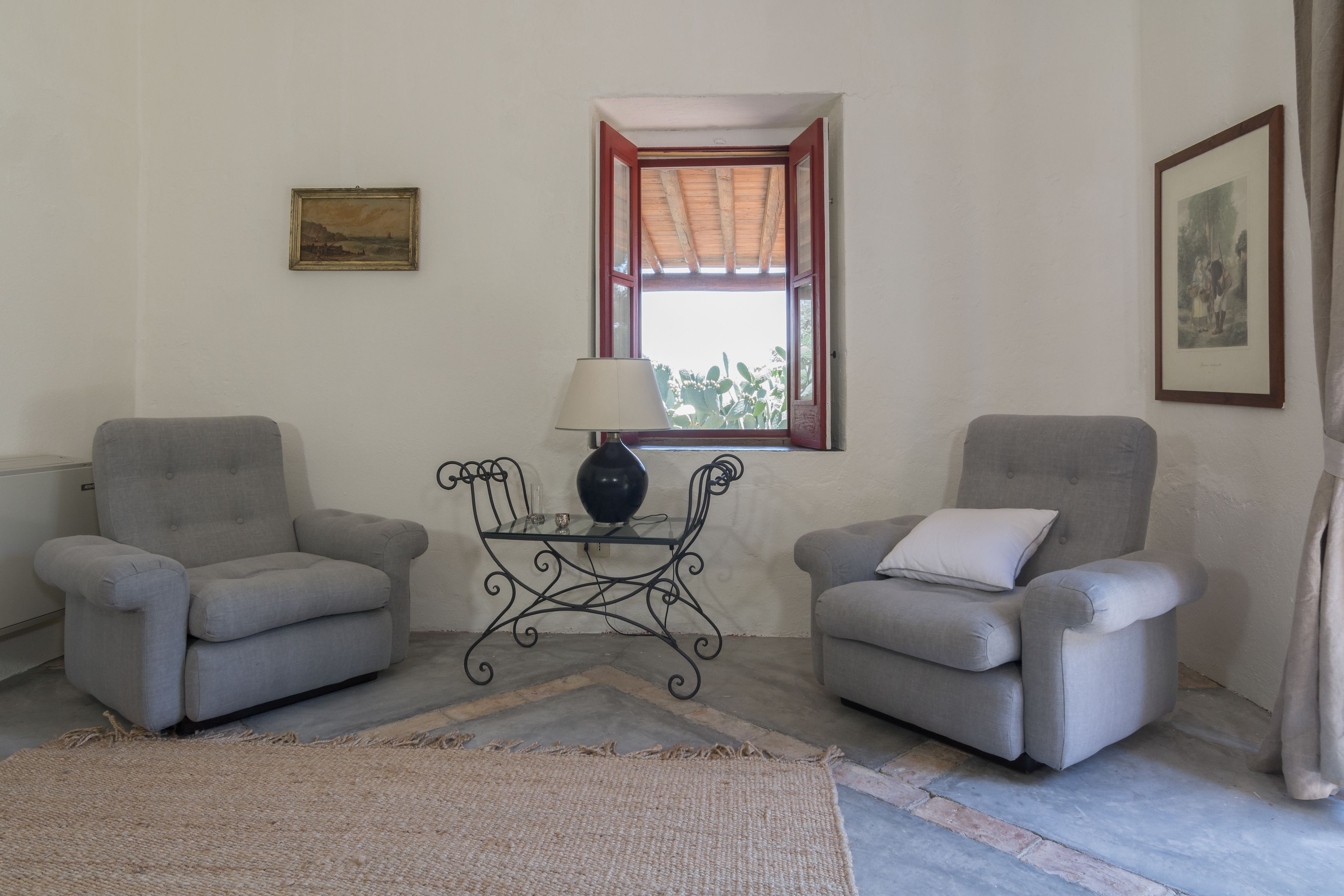 A room with two grey armchairs, a small table with a lamp, a window with a red frame, and a framed picture on the wall.