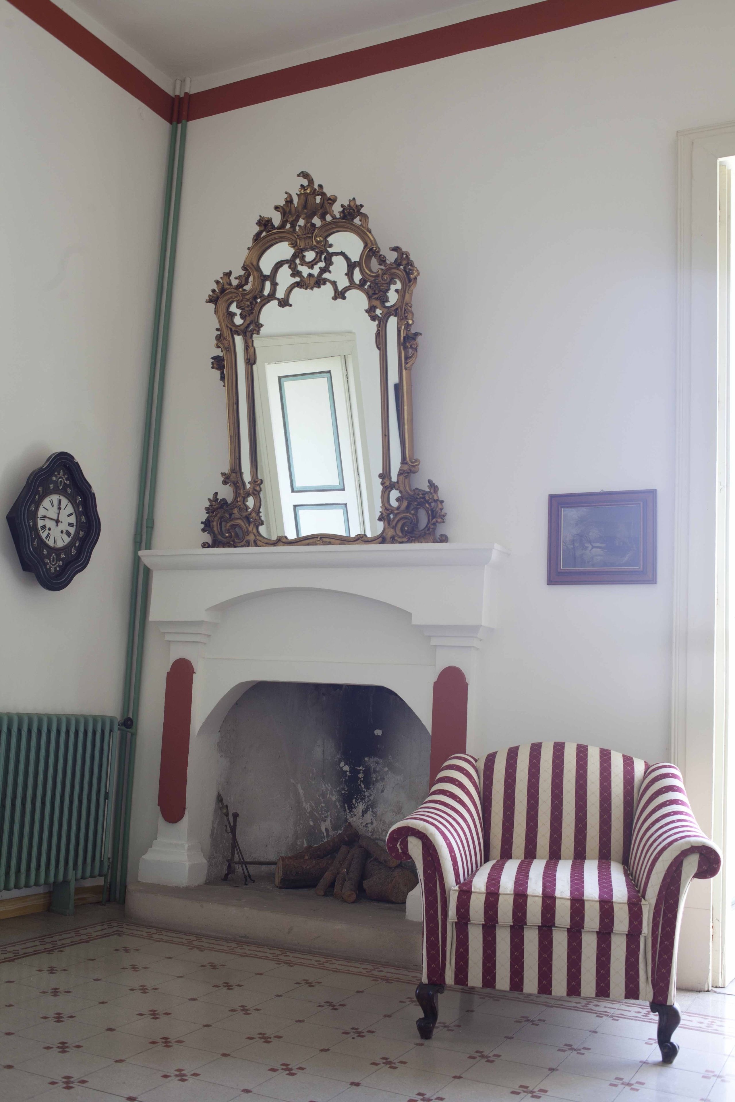 Una habitación con una chimenea blanca, un sillón a rayas rojo y blanco, un espejo grande y ornamentado, y un cuadro en la pared.
