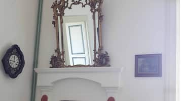 Una habitación con una chimenea blanca, un sillón a rayas rojo y blanco, un espejo grande y ornamentado, y un cuadro en la pared.