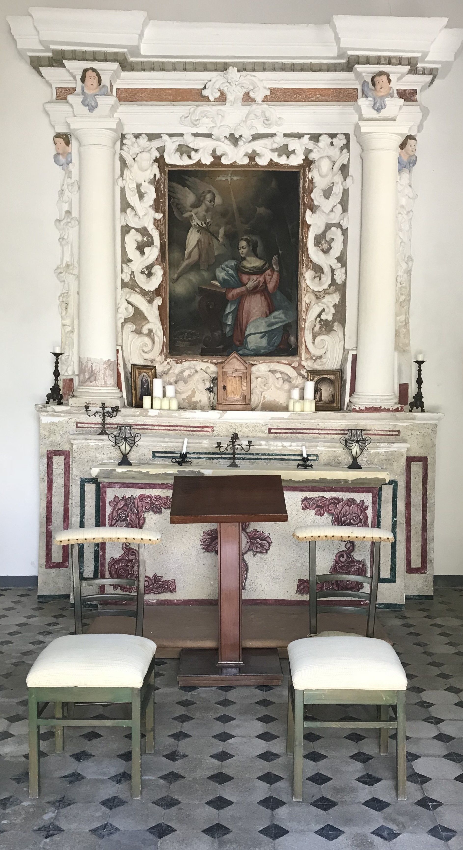 Una capilla con columnas ornamentadas, un cuadro religioso, velas y dos bancos.