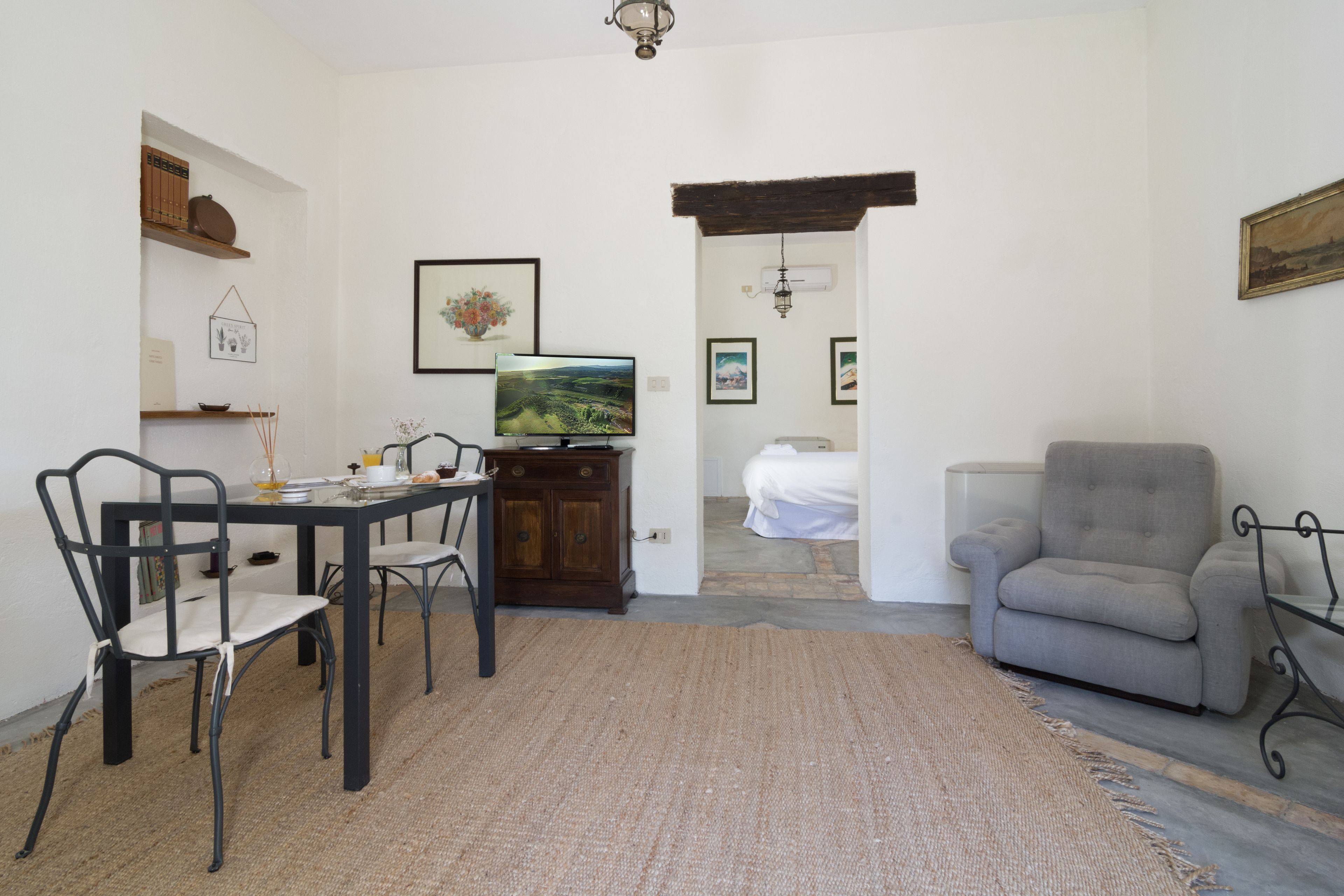 Un comedor con mesa, sillas y televisor. Se ve una cama a través de un umbral abierto.