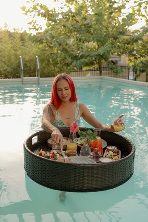Une personne assise au bord d’une piscine, avec un plateau flottant rempli de nourriture et de boissons.
