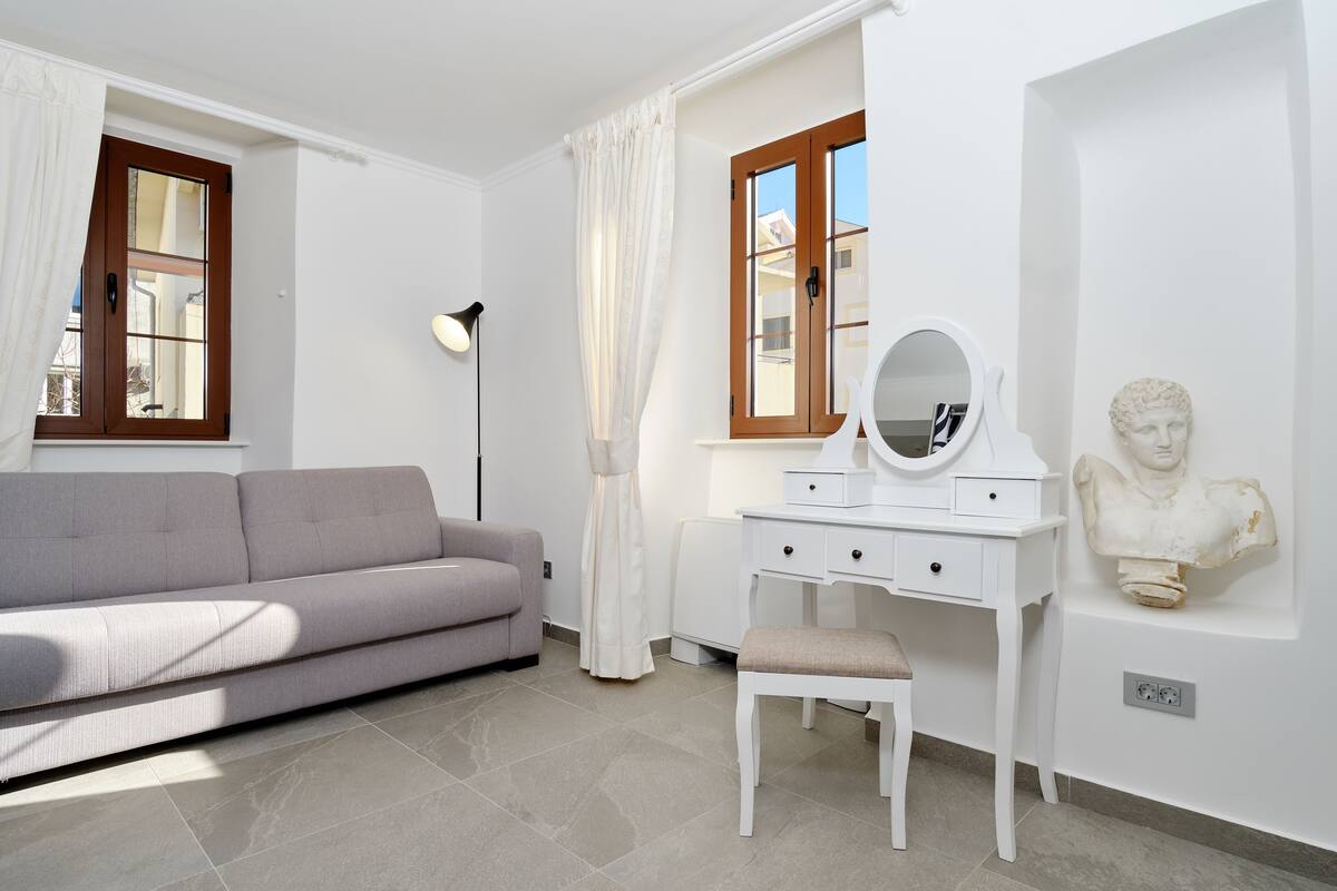 A modern bedroom with a grey sofa, a white dresser with a mirror, and a bust sculpture on the wall.
