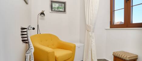 A room with a yellow armchair, a small wooden stool, a white door, a framed picture, and a window with curtains.