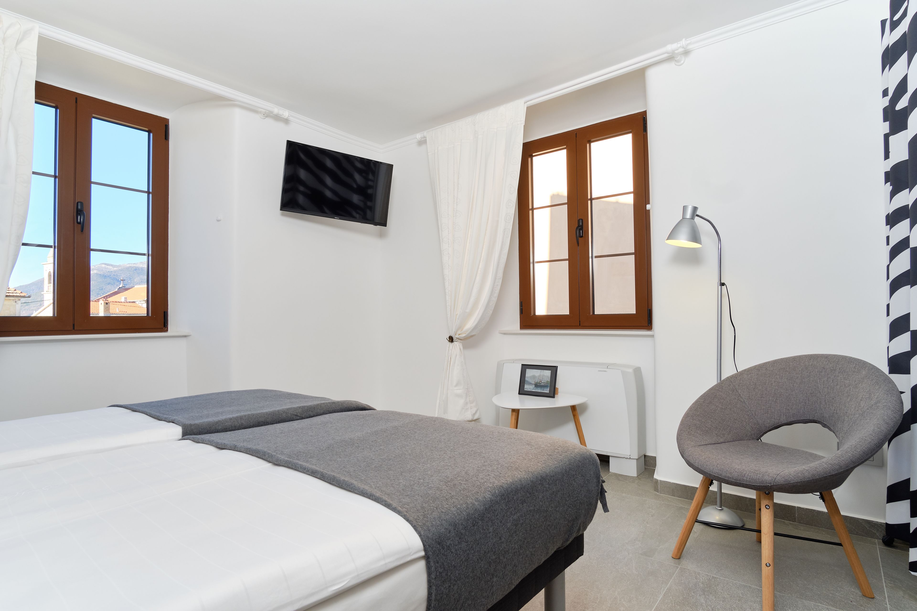 A hotel room with two beds, a small table with a picture frame, a chair, a TV mounted on the wall, and a window with curtains.